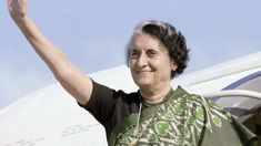 an older woman waves at the camera as she stands in front of an airplane with her hand up