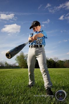 a young boy holding a baseball bat on top of a lush green field with clouds in the background