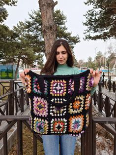 a woman holding up a crocheted blanket on top of a wooden fence next to a tree