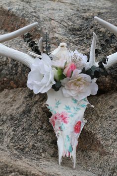 an animal skull with flowers on it's antlers sitting on the ground next to some rocks