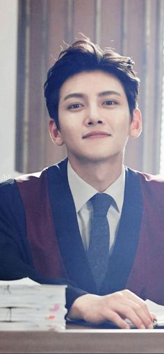 a young man in a sweater and tie sitting at a desk with stacks of papers