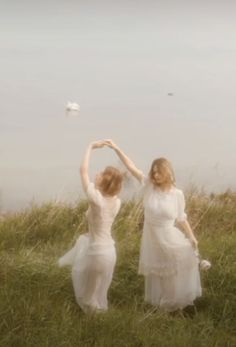 two women in white dresses are dancing on the grass next to a body of water