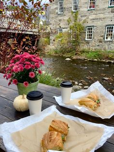 two sandwiches and coffee sit on a picnic table next to a small river with flowers in the background