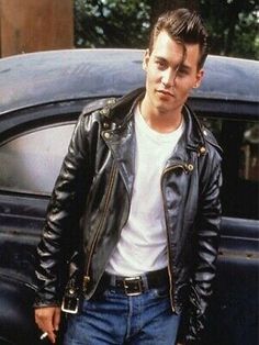 a man standing in front of a car wearing a leather jacket and jeans with his hands in his pockets