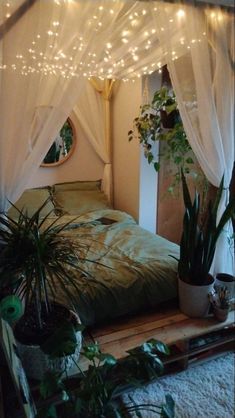 a bed with white drapes and plants in the corner next to it is lit up by fairy lights