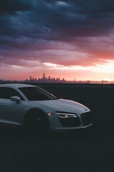 a white car parked in the middle of a field at sunset with a city skyline behind it