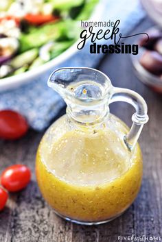 homemade greek dressing in a small glass pitcher with tomatoes and cucumbers behind it