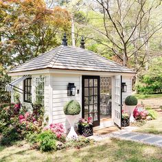 a small white shed with potted plants and flowers around the outside door is surrounded by greenery