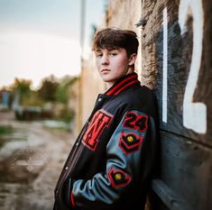 a young man leaning against the side of a building wearing a black and red jacket