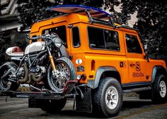 an orange vehicle with a motorcycle on the back is parked in front of some trees