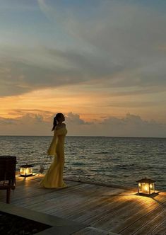 a woman in a long yellow dress standing on a dock at sunset with candles lit up