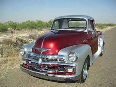 an old red and silver truck parked on the side of the road with grass in the background