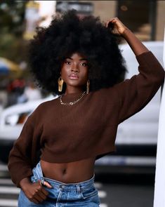 a woman with an afro standing next to a white pole wearing jeans and a brown sweater