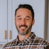 a man in plaid shirt standing in front of white cupboards and smiling at the camera