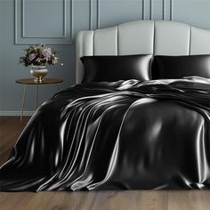 a bed covered in black satin sheets and pillows with flowers on the side table next to it