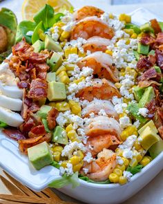 a salad with shrimp, corn, avocado and bacon on it is sitting on a white plate