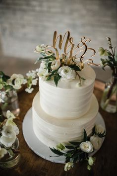 a wedding cake with white flowers and greenery on the top is sitting on a wooden table