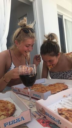 two women eating pizza and drinking wine at a table with boxes of pizza on it