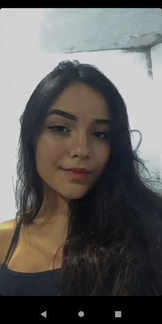 a woman with long dark hair standing in front of a white wall and looking at the camera