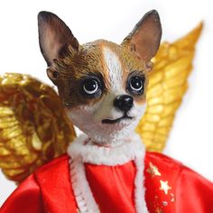 a small dog dressed in red and gold with wings on its head, sitting next to a golden angel figurine