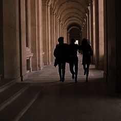 two people walking down a long hallway with columns on either side and light at the end