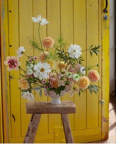 a white vase filled with lots of flowers sitting on top of a wooden stool next to a yellow door