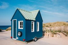 a tiny blue house with a green roof and white windows on the beach next to sand dunes