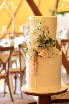 a wedding cake with white flowers and greenery sits on a table in a tent