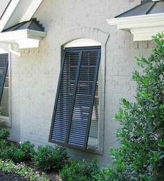 two windows with black shutters on the side of a house in front of bushes and trees