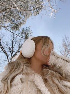 a woman with long hair wearing ear muffs and fur coat in front of trees