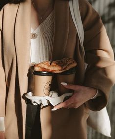 a woman holding a coffee cup and some food in her hand while wearing a brown coat