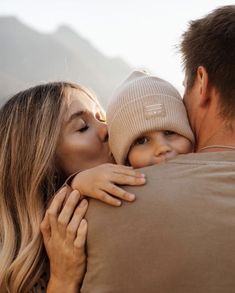 a man and woman kissing each other while holding a small child in their arms with mountains in the background