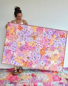 a woman holding up a large painting on top of a colorful rug in front of a white wall