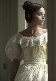 a woman in a white dress is standing by a window and looking off into the distance