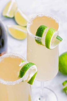 two glasses filled with lemonade and limes on top of a white tablecloth