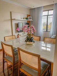 a dining room table with chairs and flowers in a vase on the centerpiece, next to a window