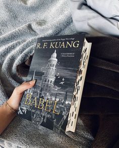 a person holding a book in their hand on top of a bed next to a blanket