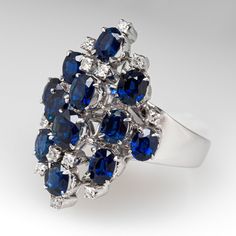 This stunning cluster style ring is accented with twelve (12) oval mixed cut natural sapphires set into four-prong settings and twelve (12) round single cut diamonds set into four-prong settings. The ring has a tapered shank. The ring measures 24.5mm at the top, rises 12.2mm above the finger, tapering to 3.1mm wide and 1.0mm thick at the base of the shank. It is currently a size 6. One of the diamond accents is chipped and unnoticeable. Luxury Multi-stone Sapphire Cluster Ring, Sapphire Multi-stone Oval Diamond Ring, Elegant Multi-stone Sapphire Cluster Ring, Oval Multi-stone Sapphire Ring In Platinum, Luxury Multi-stone Cluster Sapphire Ring, Elegant Sapphire Multi-stone Cluster Ring, Dazzling Oval Multi-stone Cluster Ring, Fine Jewelry Sapphire Cluster Ring With Prong Setting, Sapphire Multi-stone Cluster Diamond Ring