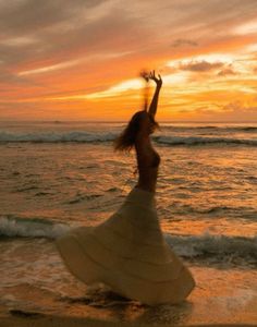 a woman standing on top of a sandy beach next to the ocean at sunset with her arms in the air