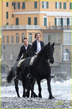 two people riding on the backs of horses in front of a building and ocean waves