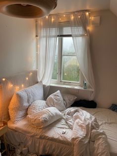 an unmade bed in front of a window with fairy lights hanging from the ceiling