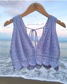 a purple crocheted top hanging on a wooden hanger at the ocean's edge