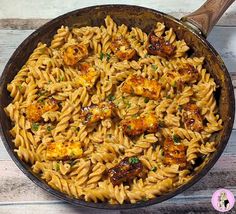 a skillet filled with pasta and meat on top of a wooden table next to a spoon