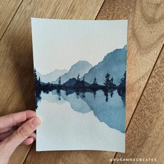 a person holding up a piece of paper with watercolor on it and mountains in the background