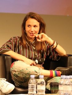 a woman sitting in a chair with her finger to her mouth and water bottles on the table behind her