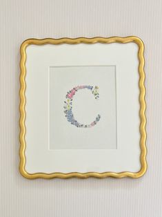 an embroidered letter c in a gold frame on a white wall with pink and yellow flowers