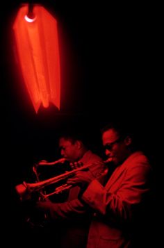 two men playing instruments in the dark with red light coming from behind them and an object hanging on the wall