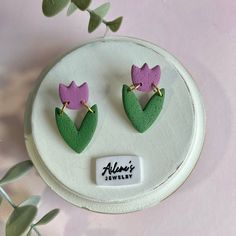 two green and purple flower shaped earrings sitting on top of a white plate next to a plant