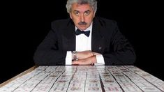 a man in a tuxedo sitting at a table with stacks of money on it