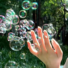 a woman blowing bubbles in the air with her hand on top of it and trees behind her